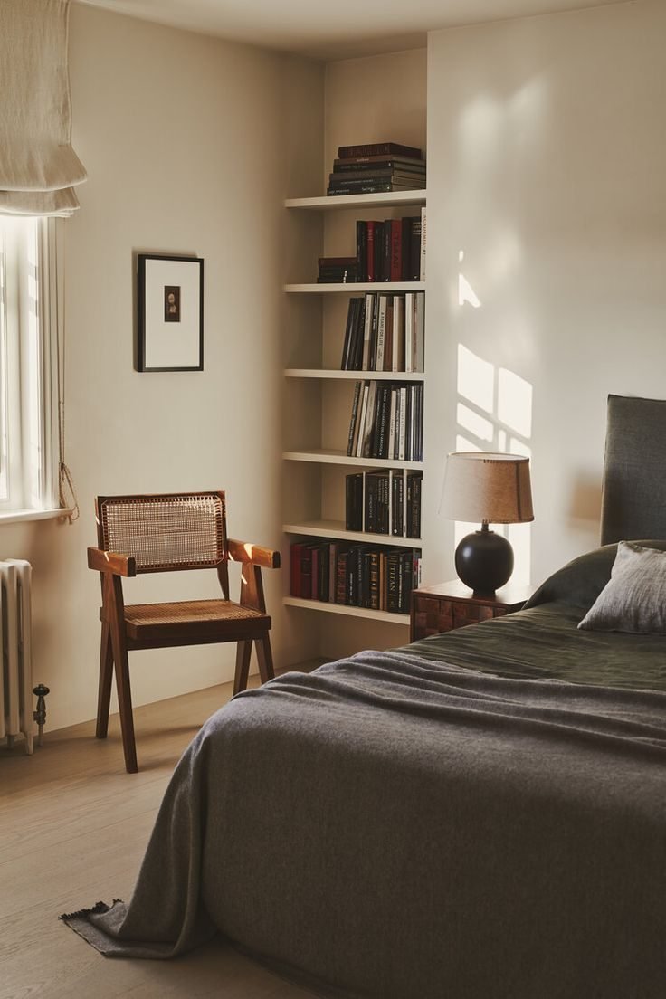 Small room: bed with a grey throw, cane armchair, built-in bookshelves and morning light.